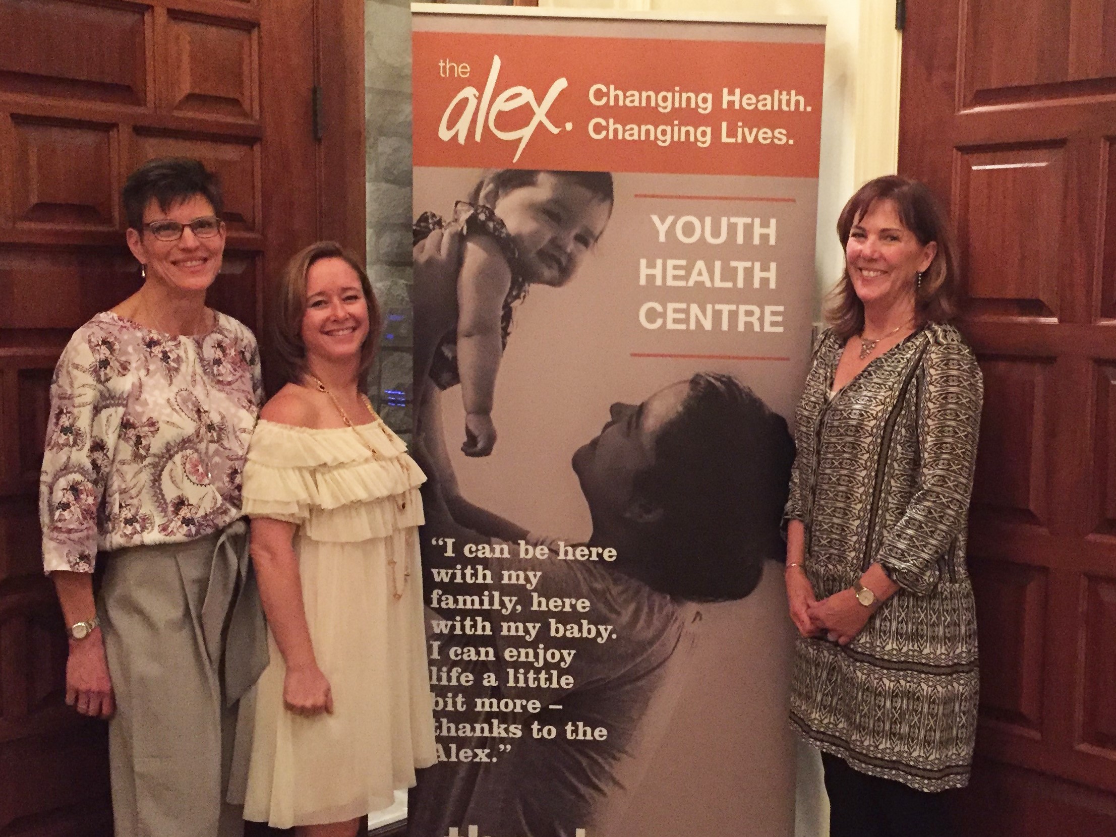 three women standing in front of an Alex Youth Health Centre banner