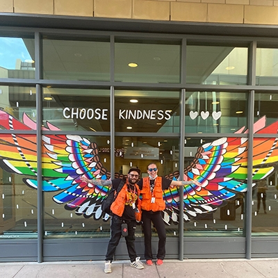 Two members of The Alex's Street Team in orange vests posing in front of a mural downtown that reads "Choose Kindness"