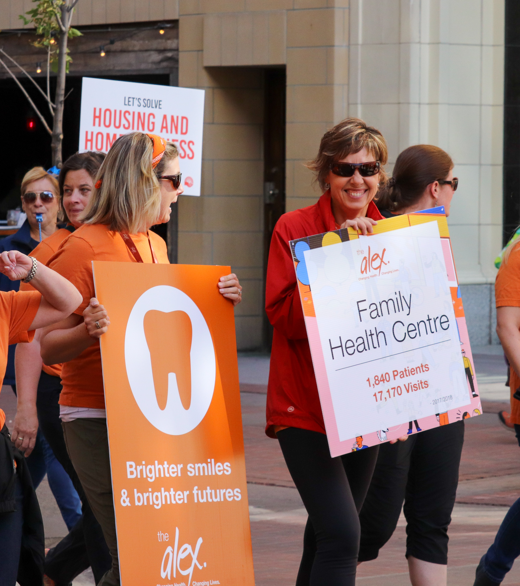 Two Alex staff walking in a parade holding signs that advertise Alex programs